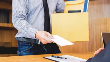 Un empleado, vestido con camisa y corbata, le da una carta de renuncia voluntaria a su jefe mientras sostiene una caja con documentos.