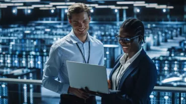 Dos personas revisan una laptop en un centro de datos iluminado.